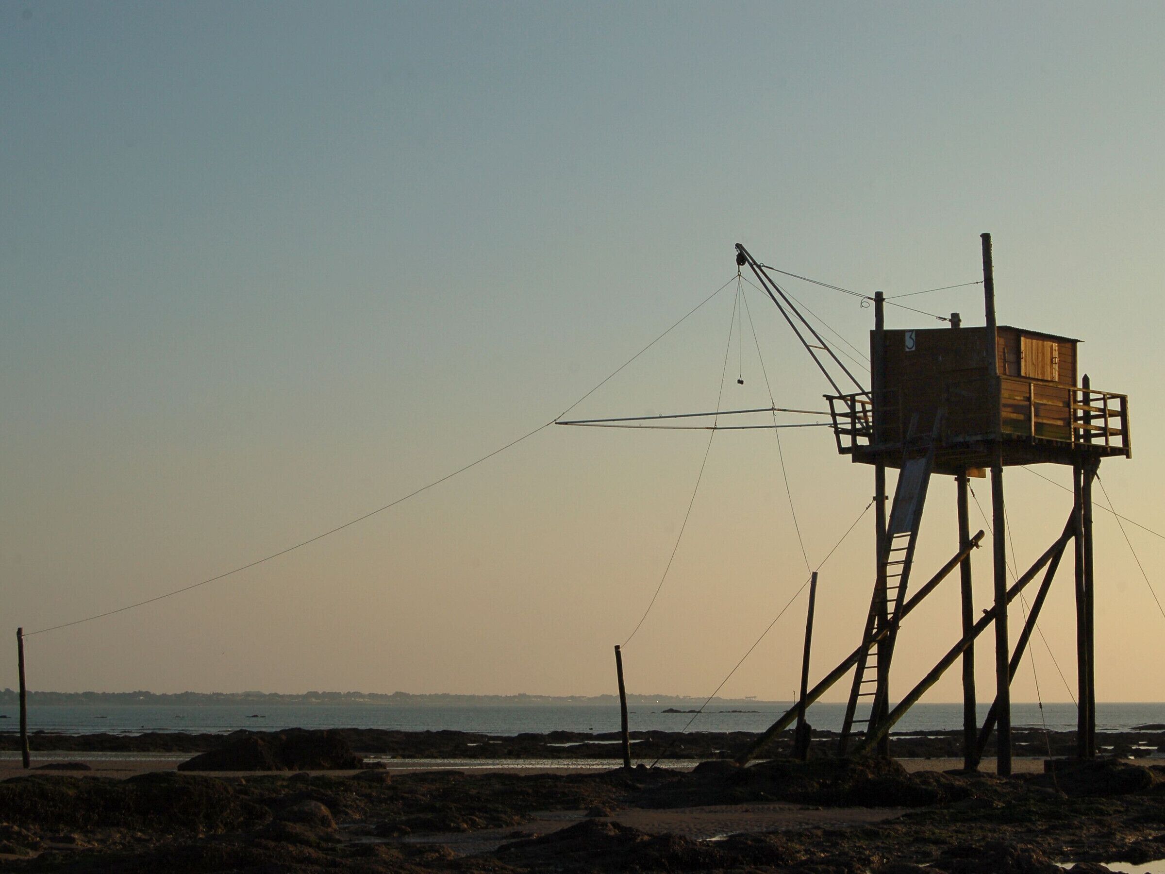 A wooden observation tower with a ladder, situated on a rocky beach with a view of the sea and distant horizon.
