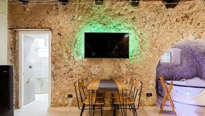 A rustic stone wall with a flat-screen TV, a wooden dining table with chairs, a bathtub, and a wooden ladder. - Antrum Loft, eaperience with Jacuzzi (Salerno)