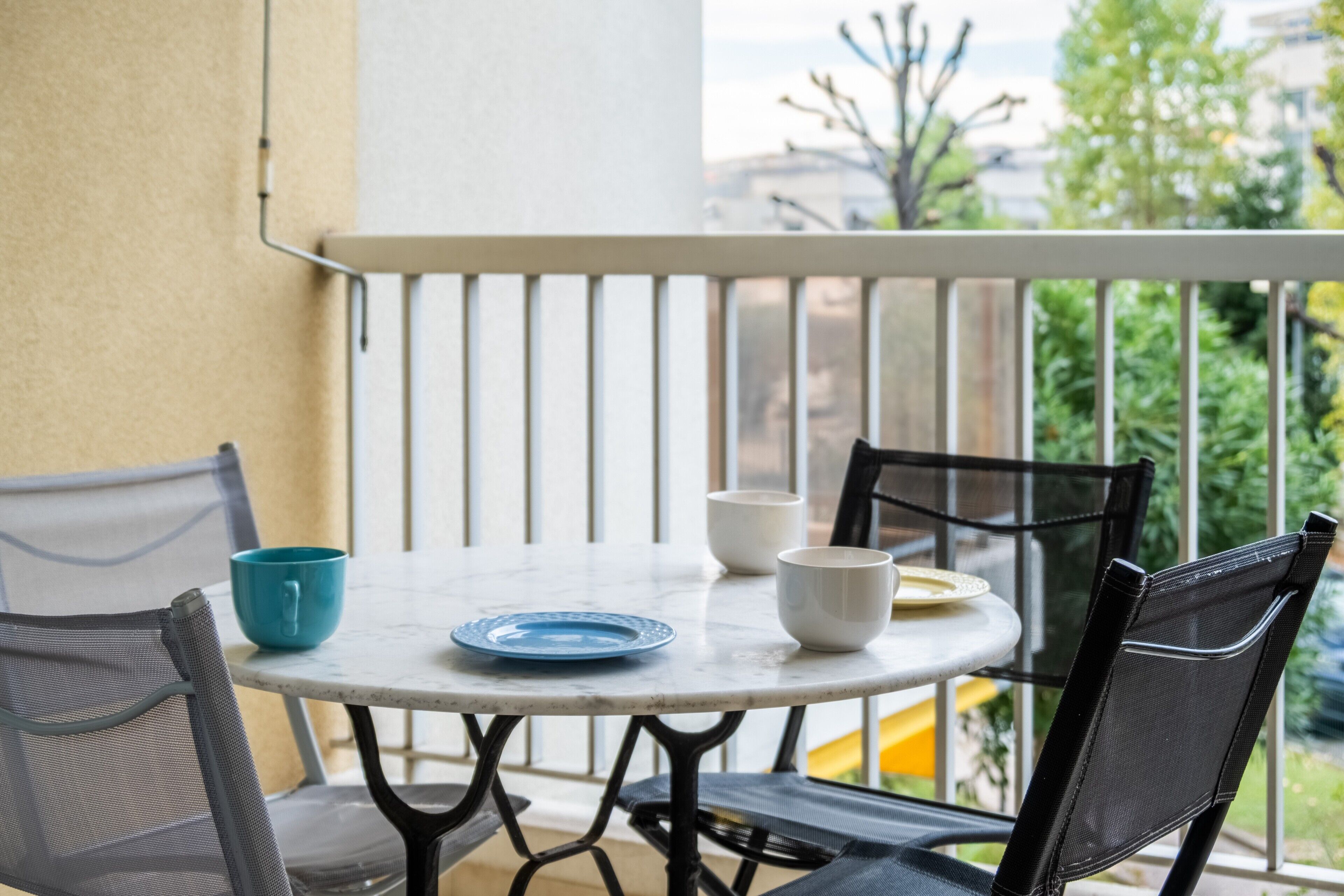 A round table with chairs, a blue plate, and white bowls on a balcony with a railing and greenery in the background.