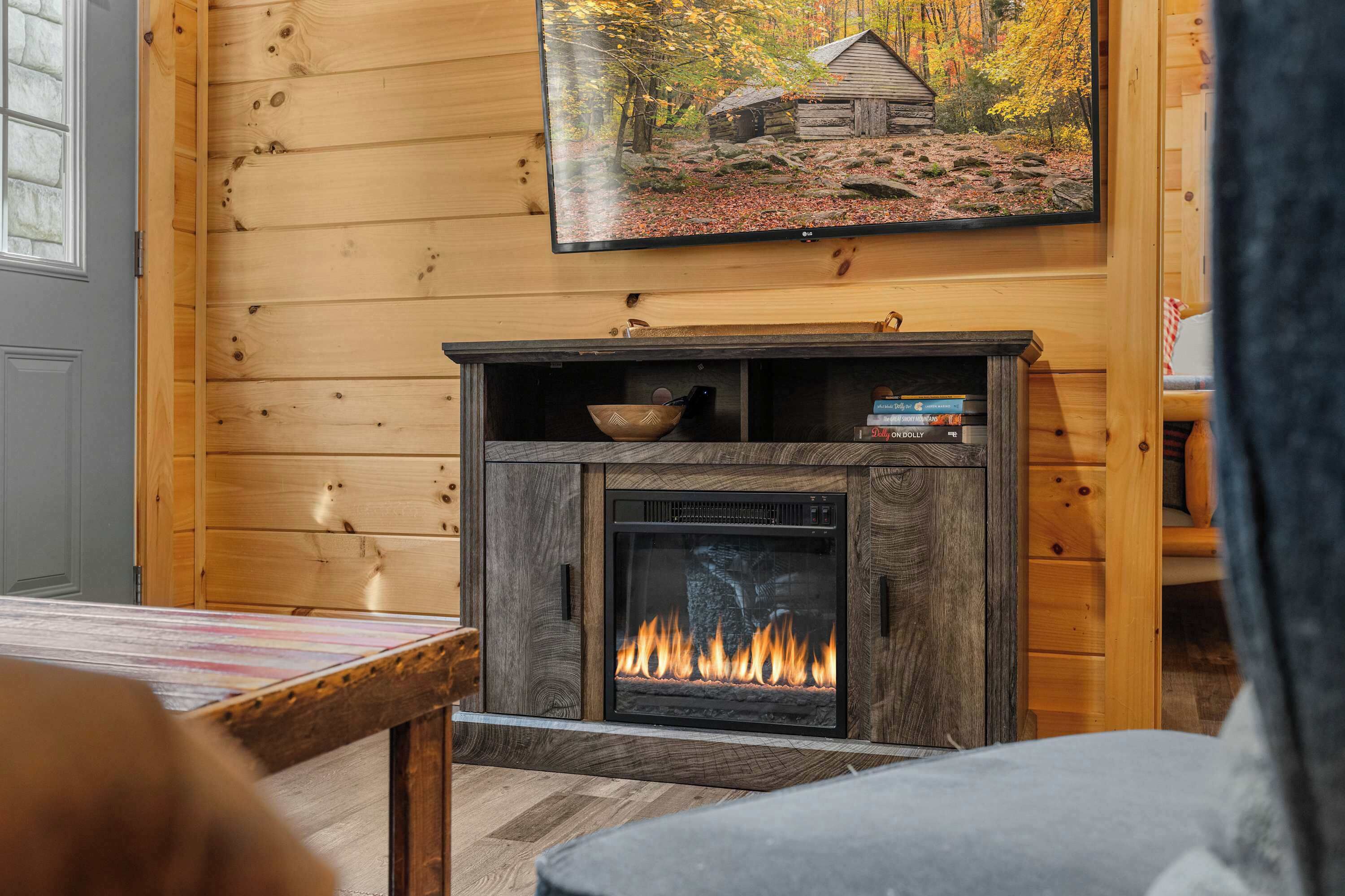A cozy room with a fireplace, a painting of a cabin in autumn, and a table with a bowl.