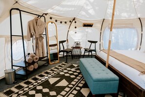 A tented room with a bed, a bench, a table with chairs, a mirror, and a rack with towels.