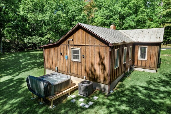 A wooden cabin with a hot tub and a small shed, surrounded by a grassy yard and trees.