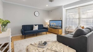 Ein modernes Wohnzimmer mit einem blauen Sofa, einem grauen Sessel, einem Couchtisch mit Büchern und einem Gesellschaftsspiel, einem Fernseher und einer Uhr an der Wand.