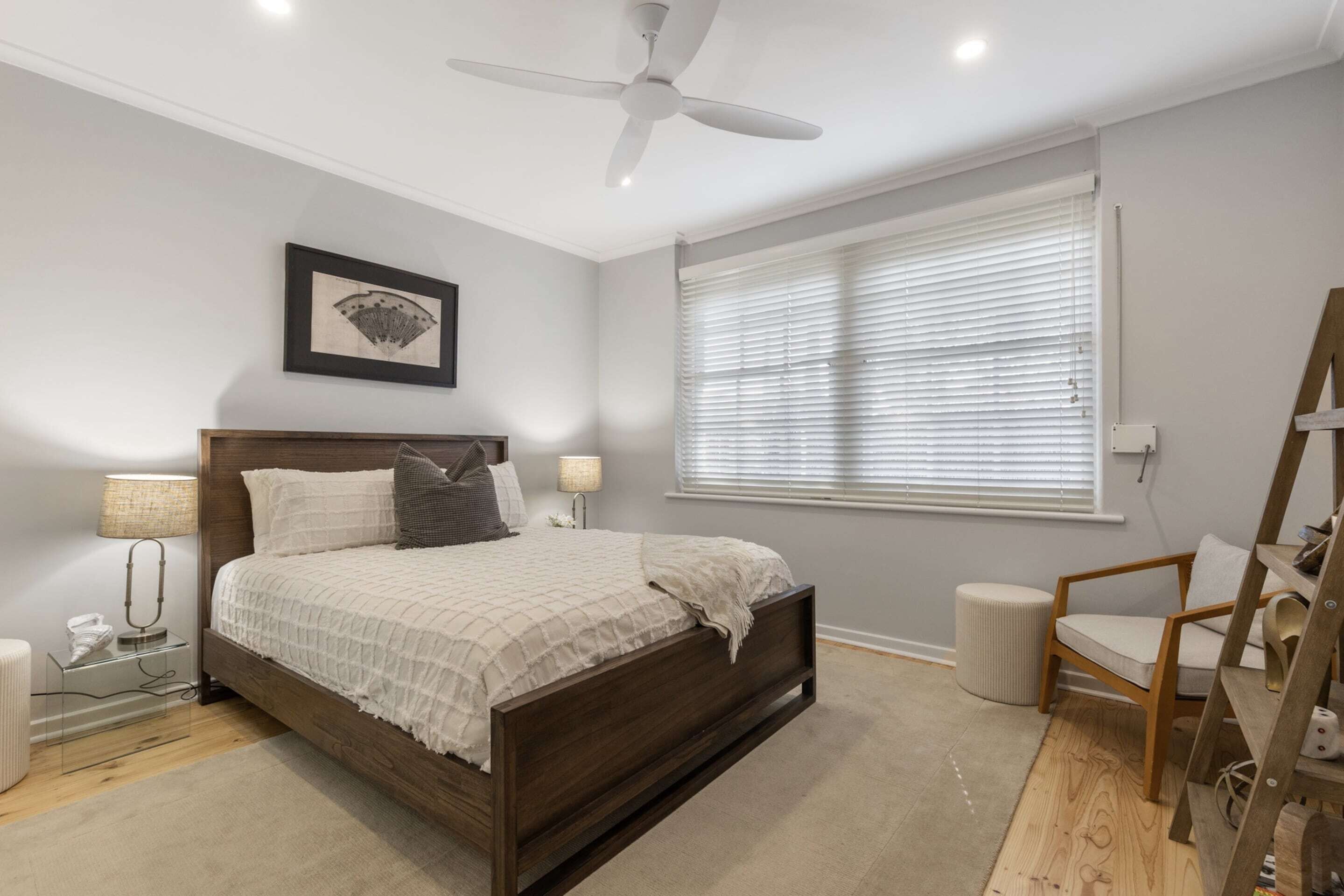 A bedroom with a wooden bed, a ceiling fan, a window with blinds, a chair, and a small table with a lamp.