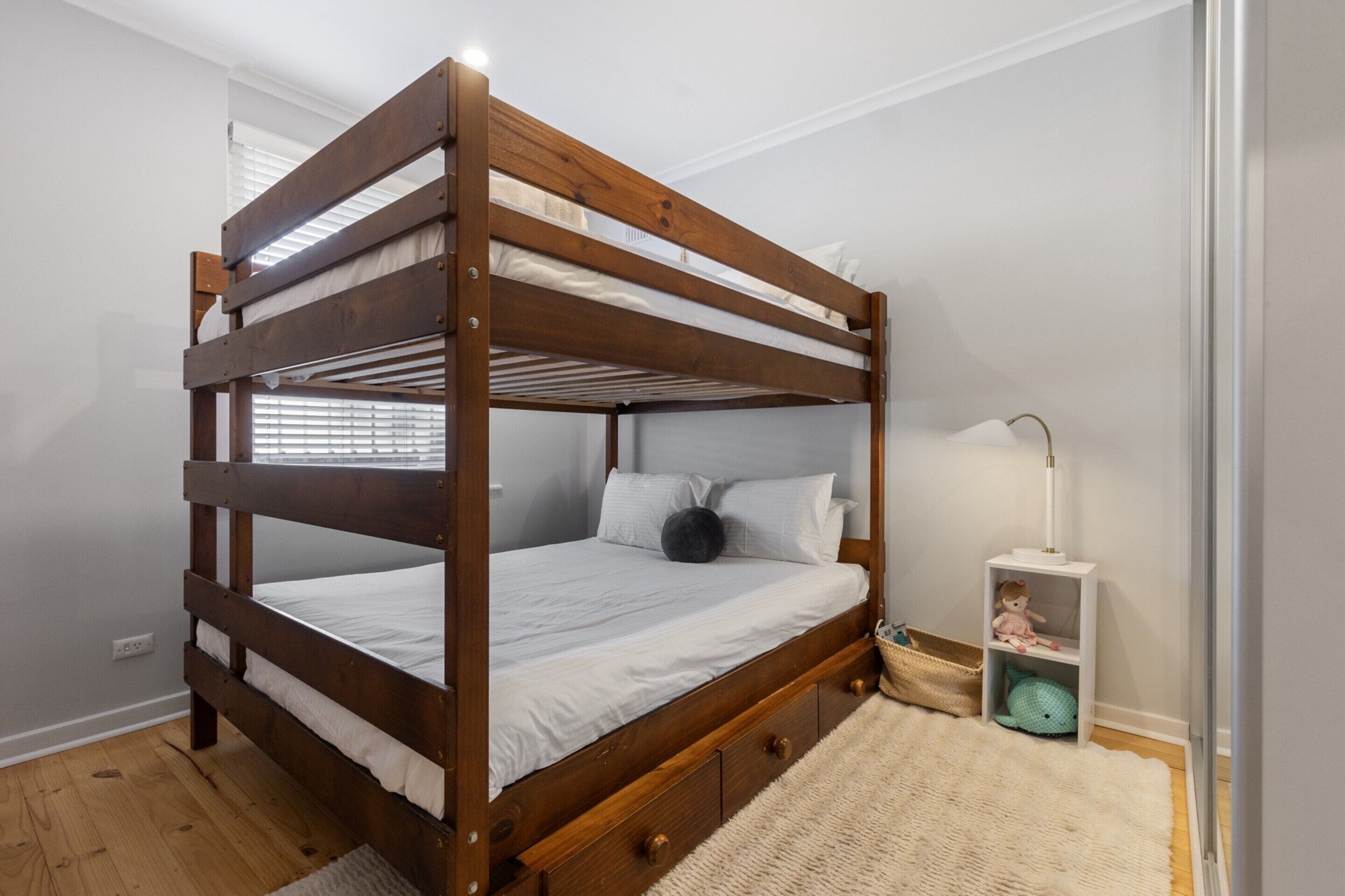 Una cama individual con estructura de madera, ropa de cama blanca, una mesita de noche con lámpara y una cesta en el suelo.