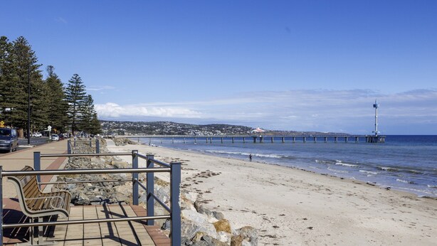 Ein Strand mit einem Pier, der ins Wasser ragt, einer felsigen Küstenlinie und einem Sitzbereich mit Geländer.