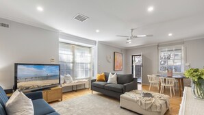 Ein modernes Wohnzimmer mit Flachbildfernseher, grauem Sofa, Hocker und einem Essbereich mit Tisch und Stühlen.