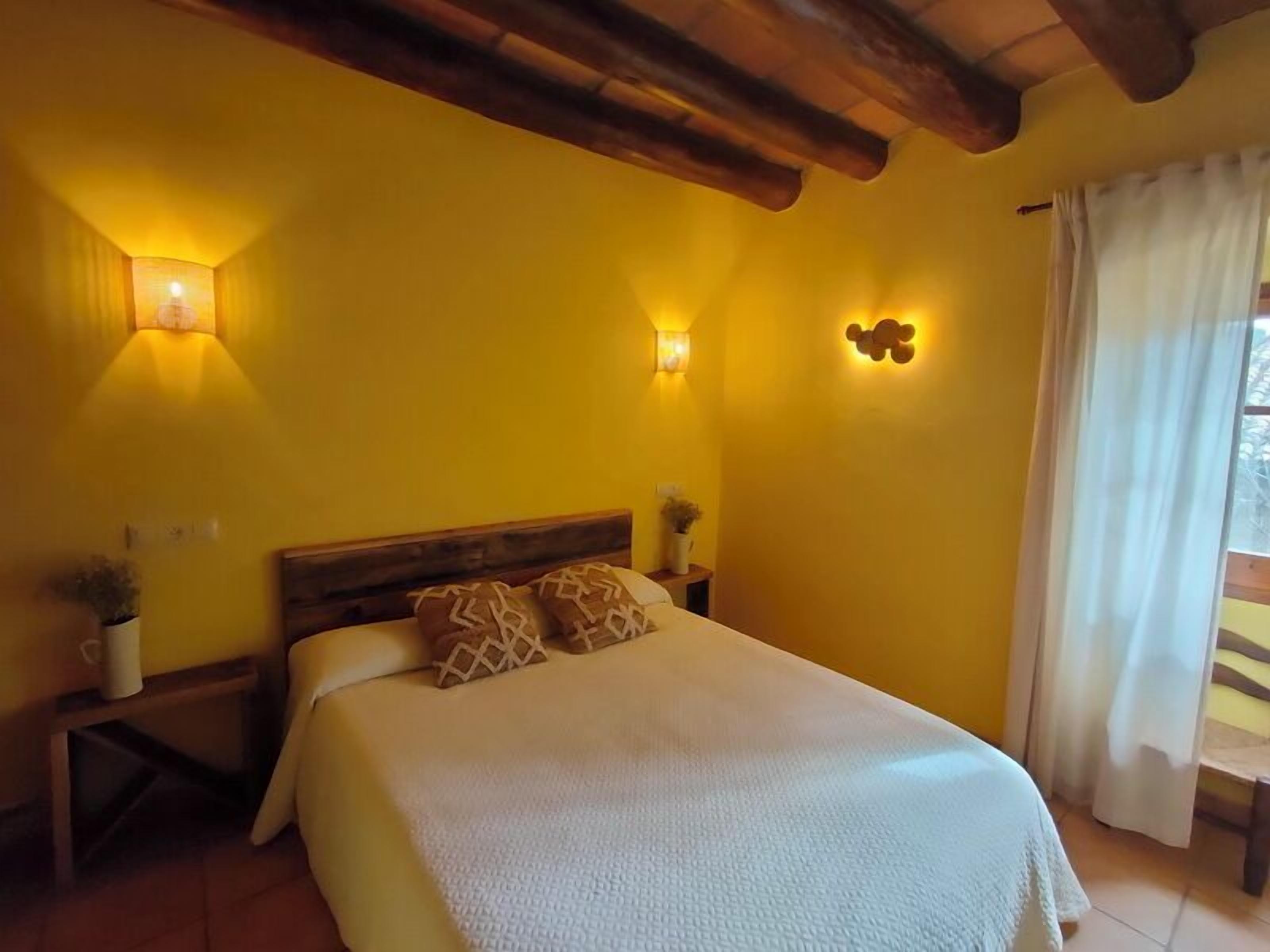 A bedroom with a wooden ceiling, a bed with a white cover, two bedside tables with lamps, and a window with curtains.