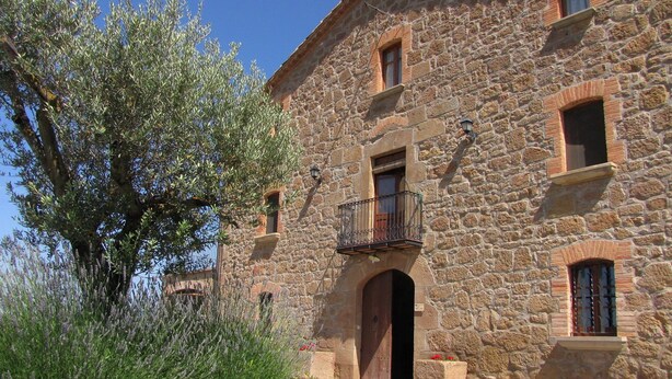 A stone building with a central entrance, a balcony, and a small garden area with lavender plants. - Casa Serra de Dalt (Lladurs)