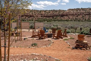 Un área de estar al aire libre con sillas de madera y una mesa, rodeada por un jardín con rocas y vegetación escasa.