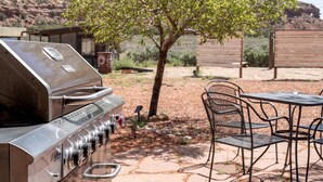 Una parrilla de acero inoxidable, mobiliario exterior con sillas y una mesa, y un árbol en un área ajardinada.