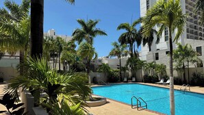 A swimming pool surrounded by palm trees and lounge chairs, with a building in the background. - Cozy Escape | Just Steps from the Beach w. Pool (Carolina)