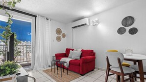 A modern living room with a red sofa, a dining table with chairs, a small coffee table, and a view of the city through a large window. - Cozy Escape | Just Steps from the Beach w. Pool (Carolina)