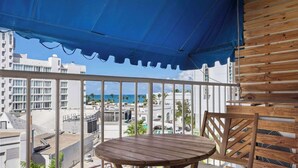 A wooden table and chair set under a blue awning on a balcony with a view of the ocean and buildings. - Cozy Escape | Just Steps from the Beach w. Pool (Carolina)