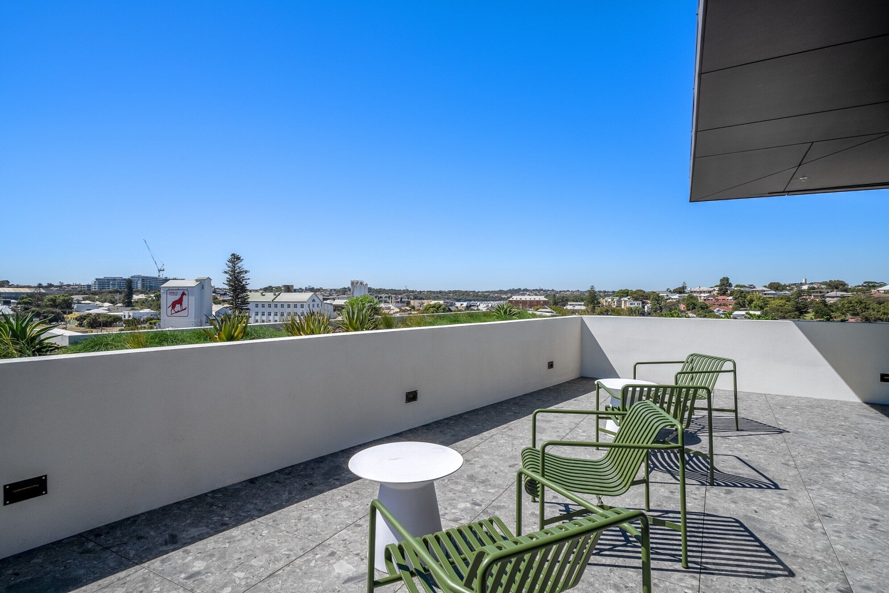 Une terrasse sur le toit, avec une table blanche et des chaises vertes, d’où l’on a une vue sur le paysage urbain.