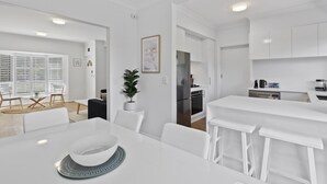 A modern kitchen with white cabinetry, a dining area with a white table and chairs, and a small bar area with stools. - Beachside 3-bed Retreat With BBQ & Garage (Adelaide)