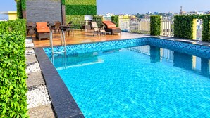 Piscina no terraço com espreguiçadeiras e área de estar, cercada por vegetação e vista para a cidade.