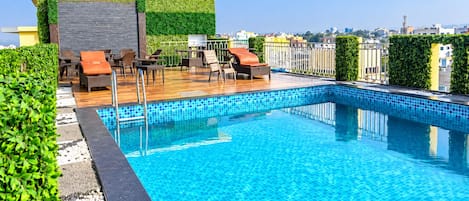 A rooftop swimming pool with lounge chairs and a seating area, surrounded by greenery and cityscape views.