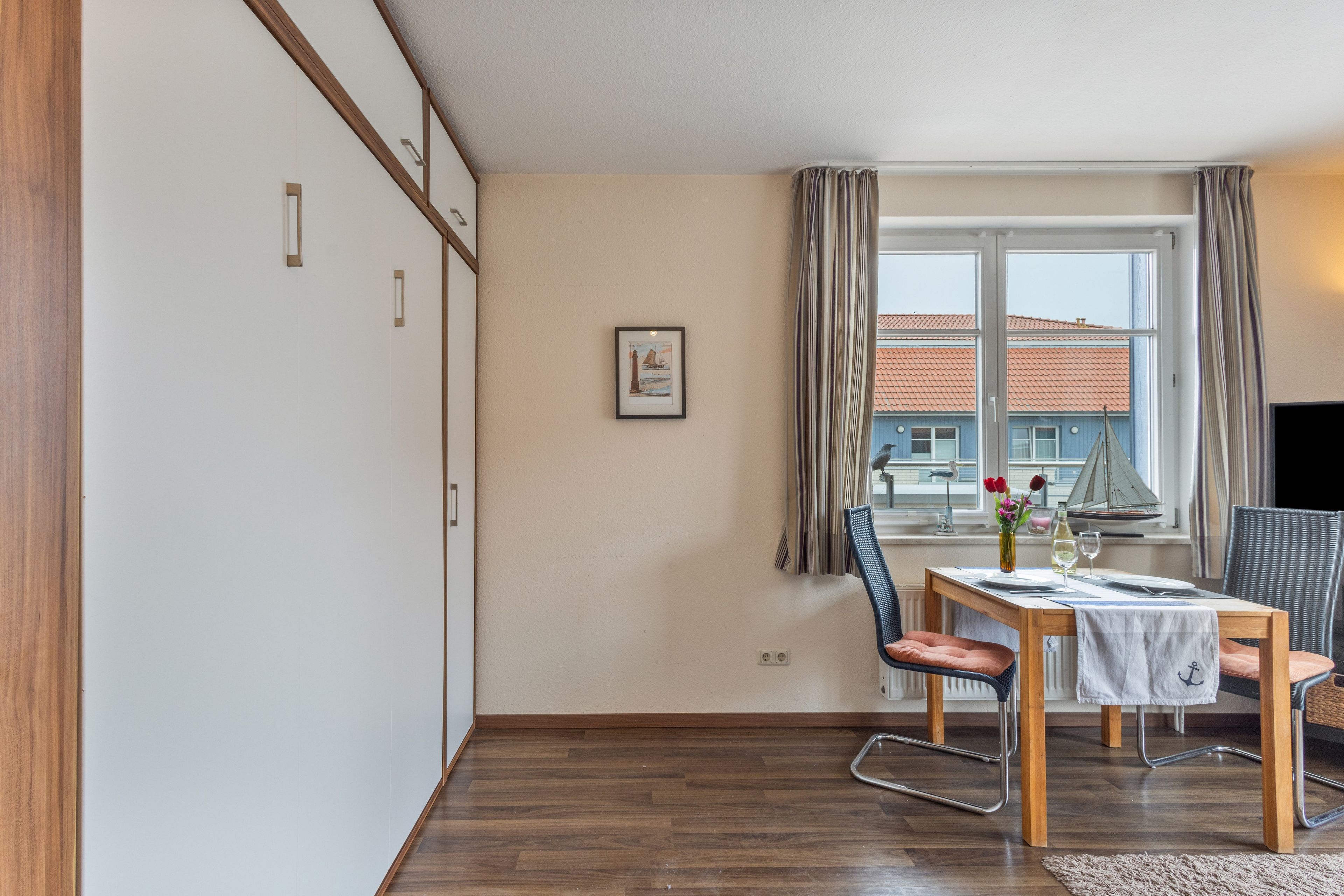Ein Zimmer mit Esstisch, Stühlen, einem Fenster mit Vorhängen, einem gerahmten Bild an der Wand und Blick auf Gebäude.