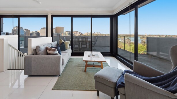 Ein modernes Wohnzimmer mit einer Couch, einem Sessel und einem Couchtisch, großen Fenstern mit Stadtblick und einem Balkon mit Gartenmöbeln.