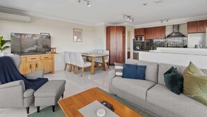 Ein modernes Wohnzimmer mit Sofa, Couchtisch und Fernseher. Es gibt eine Essecke mit Stühlen und einem Tisch. Die Küche ist sichtbar, mit Holzschränken und Geräten.
