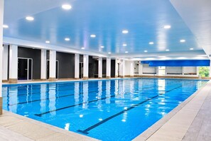 Une vaste piscine intérieure avec des couloirs bien délimités, entourée de piliers blancs et surmontée d’un plafond bleu.