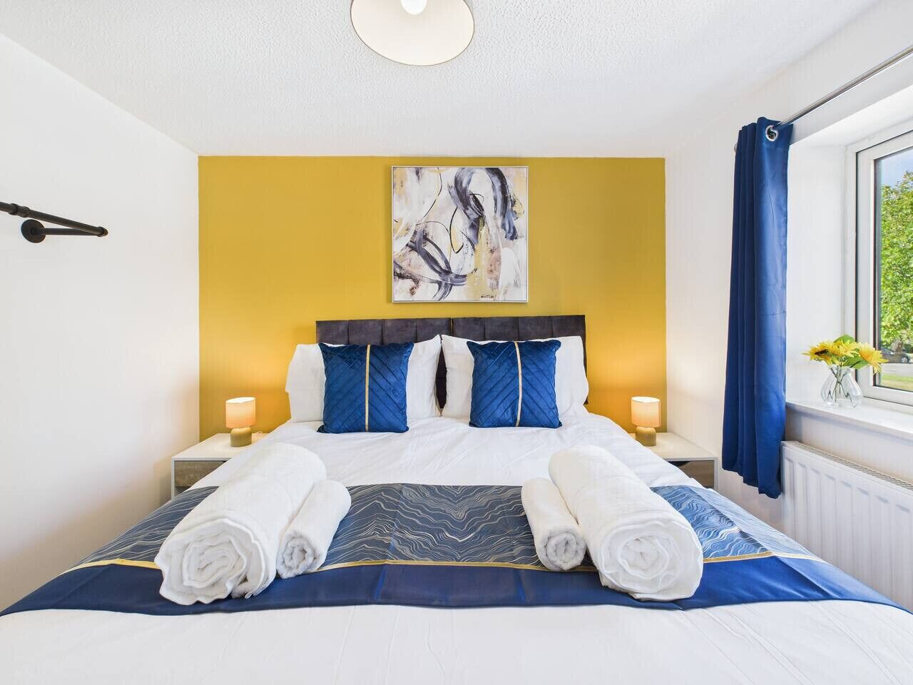 A hotel room with a bed, two bedside lamps, a window with curtains, and a painting on the wall.