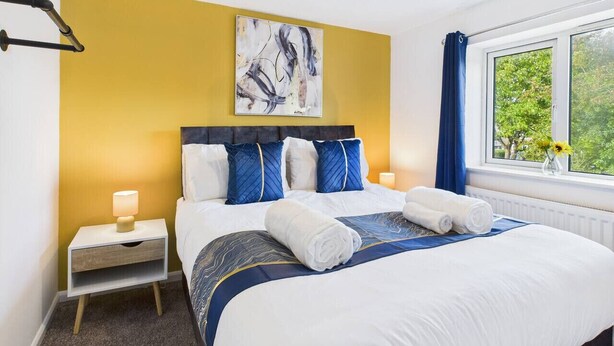 A hotel room with a bed, a nightstand, a lamp, a window with curtains, and a painting on the wall. - NEW | Cornfields Place | Racecourse | Free Parking (Bishops Cleeve)