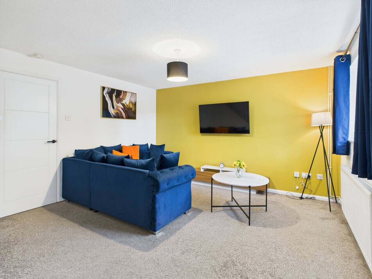 Een kamer met een blauwe bank, een klein rond tafeltje, een flatscreen televisie en een lamp.