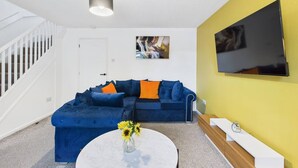 A modern living room with a blue sectional sofa, a white coffee table with sunflowers, a mounted flat-screen TV, and a staircase on the left. - NEW | Cornfields Place | Racecourse | Free Parking (Bishops Cleeve)