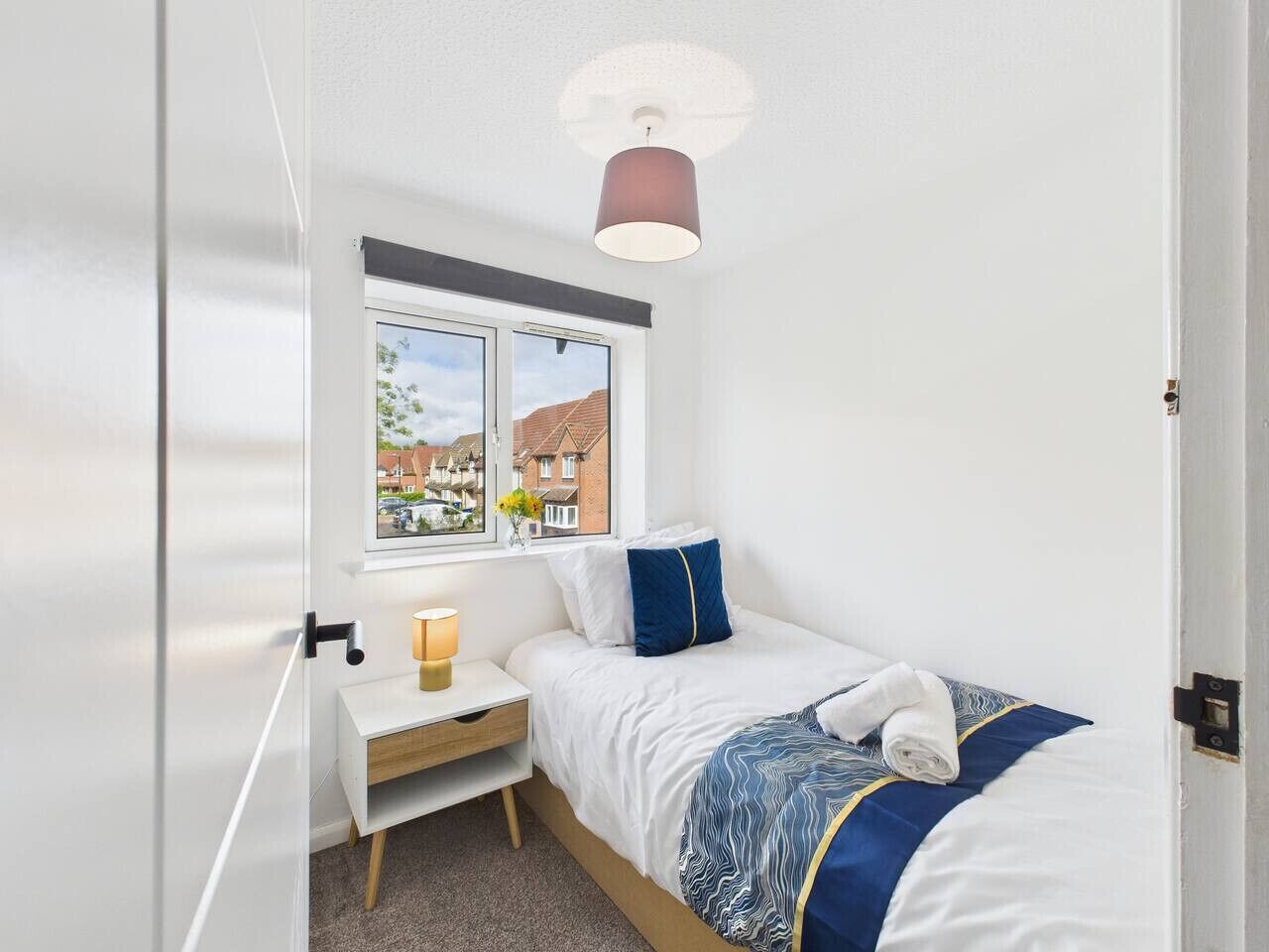 Een slaapkamer met een bed, nachtkastje, lamp, raam met uitzicht op gebouwen, en een plafondlamp.