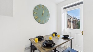 A dining area with a round wall clock, a table set with plates, glasses, and juice, and a bench. - NEW | Cornfields Place | Racecourse | Free Parking (Bishops Cleeve)