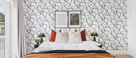 A modern bedroom with a geometric-patterned wall, a bed with a dark and orange blanket, two bedside lamps, and a small plant on the nightstand.