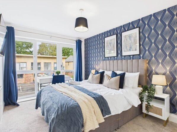 A bedroom with a large bed, a bedside table with a lamp, a window with curtains, and a patterned wall.