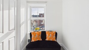 Ein kleiner Raum mit weißen Wänden, einem schwarzen Sofa und zwei orangenen Kissen mit Muster.
