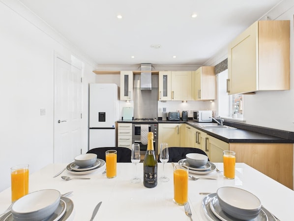 A modern kitchen with white cabinets, a central island, and a dining area set for breakfast with orange juice and a bottle of champagne.