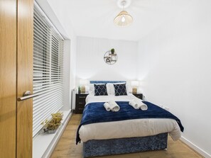 A bedroom with a bed, bedside tables, a window with blinds, a hanging light fixture, and a small shelf with a plant.