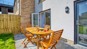 A wooden outdoor dining table with chairs, a glass door, and a brick wall. - Harvey’s Yard | Modern Coastal Home | Sleeps 6 (Cornwall)