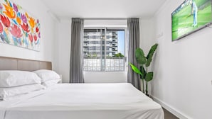 Habitación de hotel con una cama grande, ventana con vista a un edificio, un cuadro en la pared y una televisión.