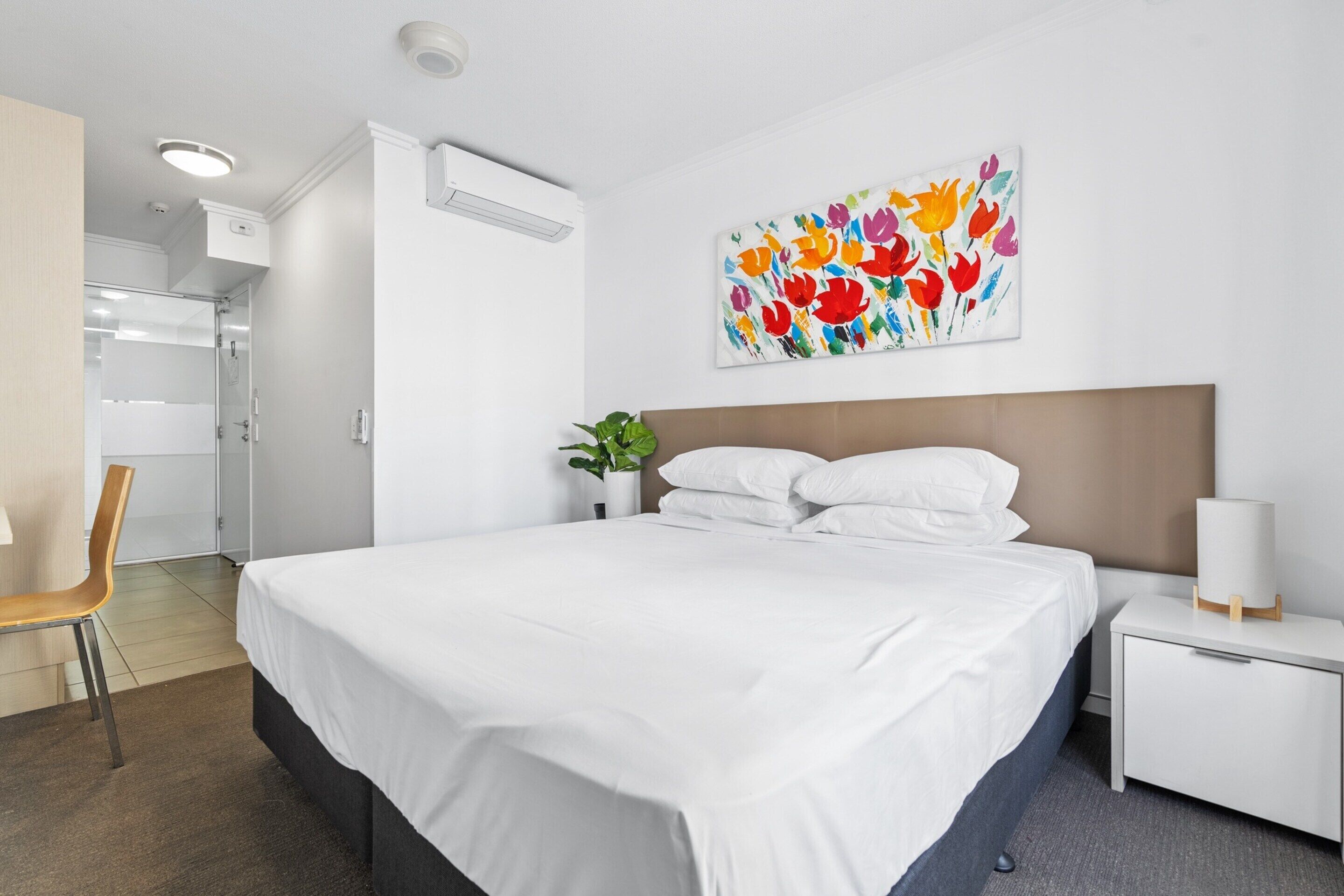 A hotel room with a large bed, a bedside table with a lamp, a wall-mounted air conditioner, and a colorful floral painting.