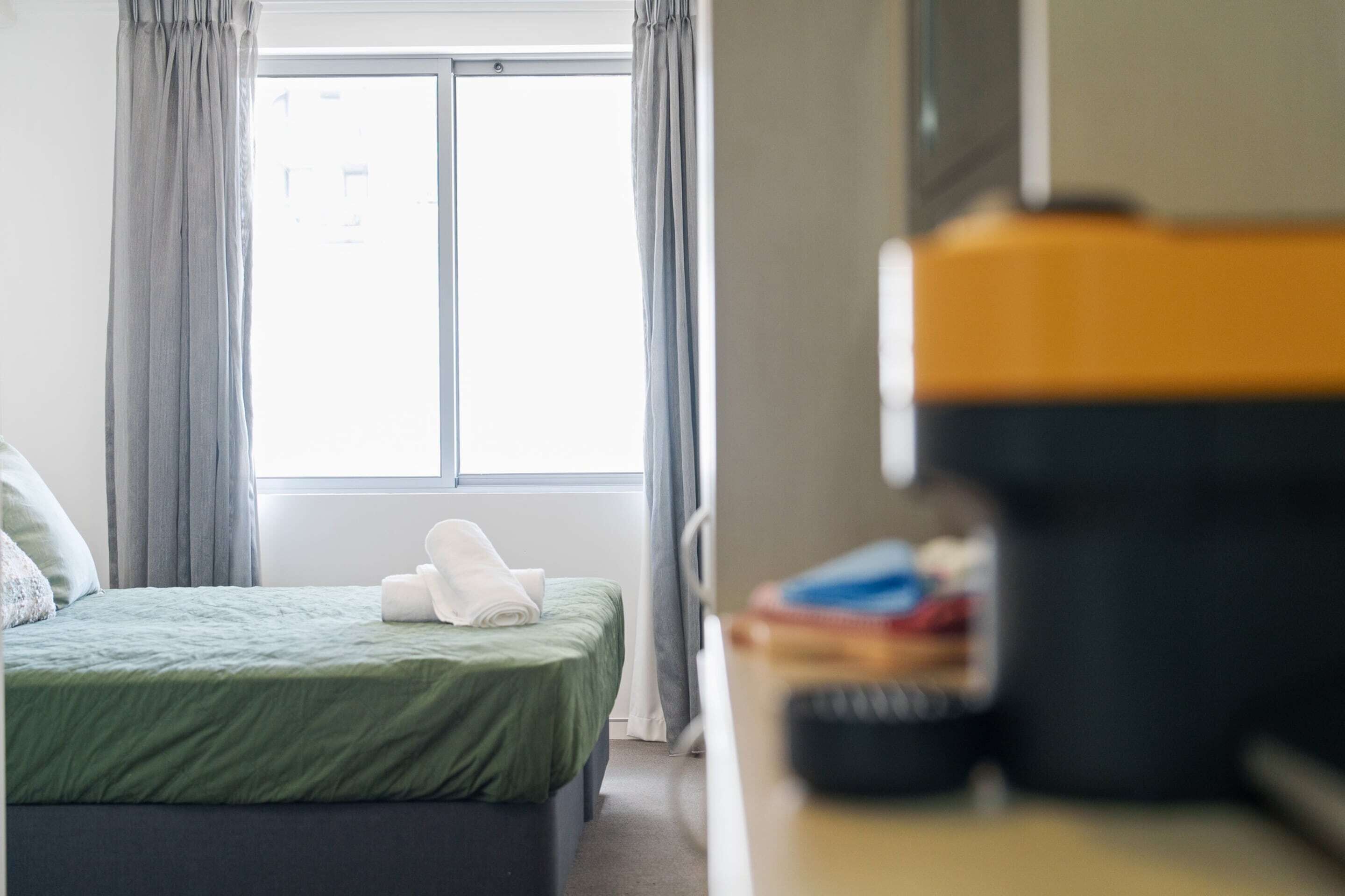 A hotel room with a bed, a window, and a desk with a computer and some books.