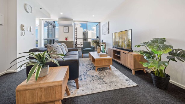 A modern living room with a black leather sofa, wooden coffee table, and a large flat-screen TV. There are potted plants and a large window providing a view of the city. - Glorious Central Canberra 1-bed With Pool & Sauna (Canberra)