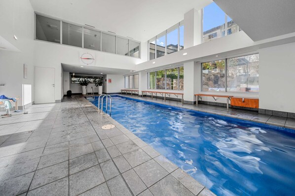 An indoor swimming pool with clear blue water, tiled floor, and benches along the sides.