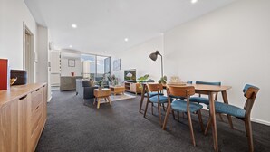 A modern living room with a dining area, featuring a wooden dining table with blue chairs, a small side table, and a lamp. - Glorious Central Canberra 1-bed With Pool & Sauna (Canberra)