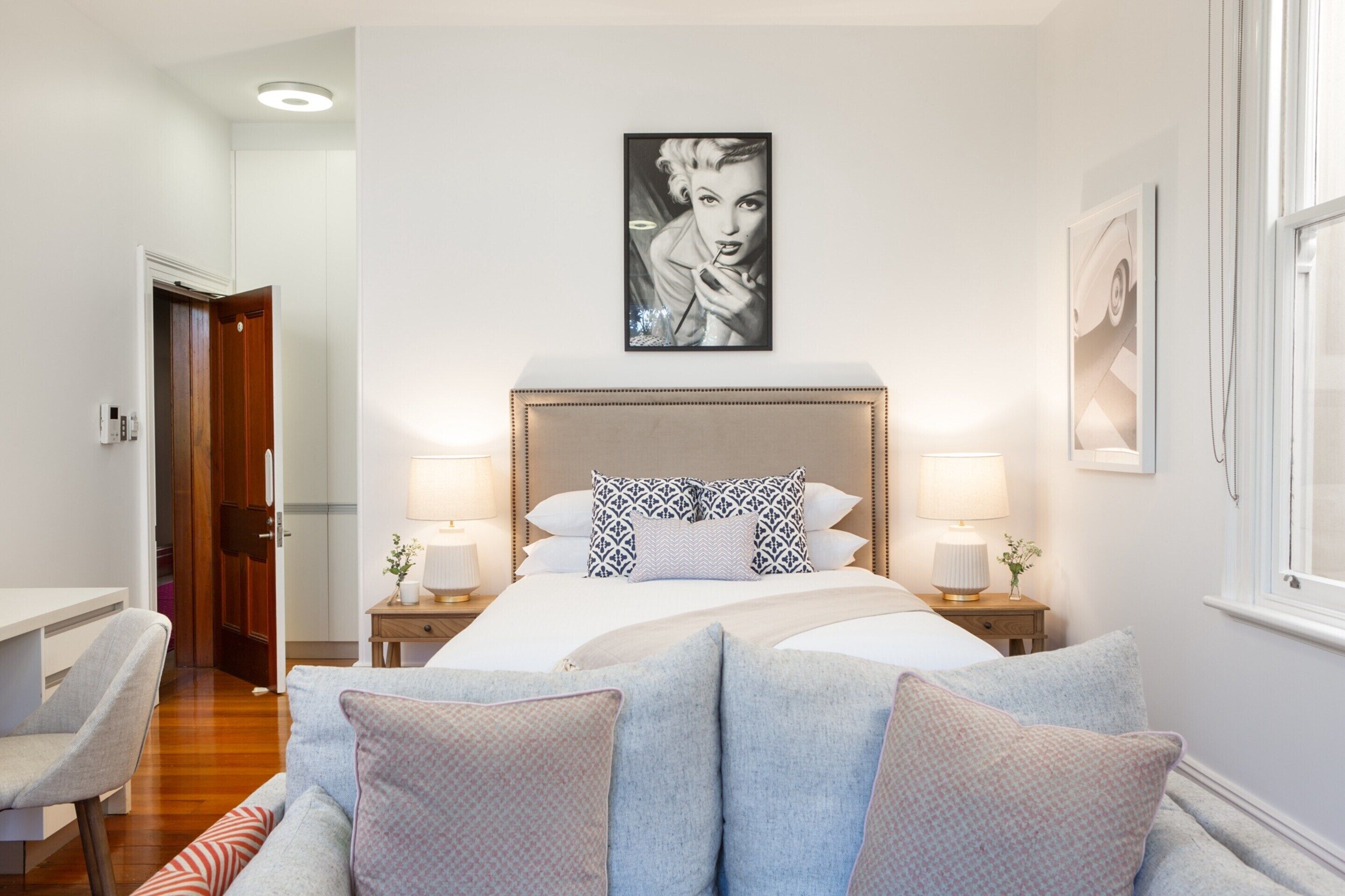 Une chambre avec un grand lit, deux tables de chevet avec des lampes, une chaise, une porte en bois et un tableau encadré au mur.