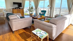 A spacious living room with a large window offering a view of the sea, a television, and a cozy seating area with a plant and a small table.