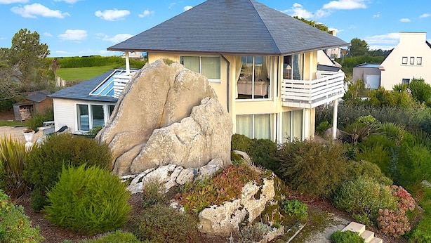 A modern house with a large rock in the garden, a balcony, and a view of the surrounding landscape.