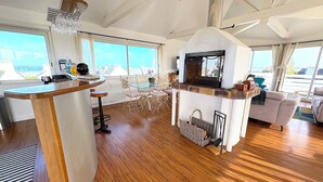 A spacious living area with a wooden floor, a central fireplace, a bar area with stools, and large windows offering a view of the sea.
