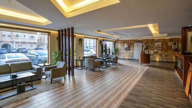 Un hall d’hôtel moderne avec un plancher en bois, des coins salon et de grandes fenêtres donnant sur les voitures garées et les bâtiments environnants.