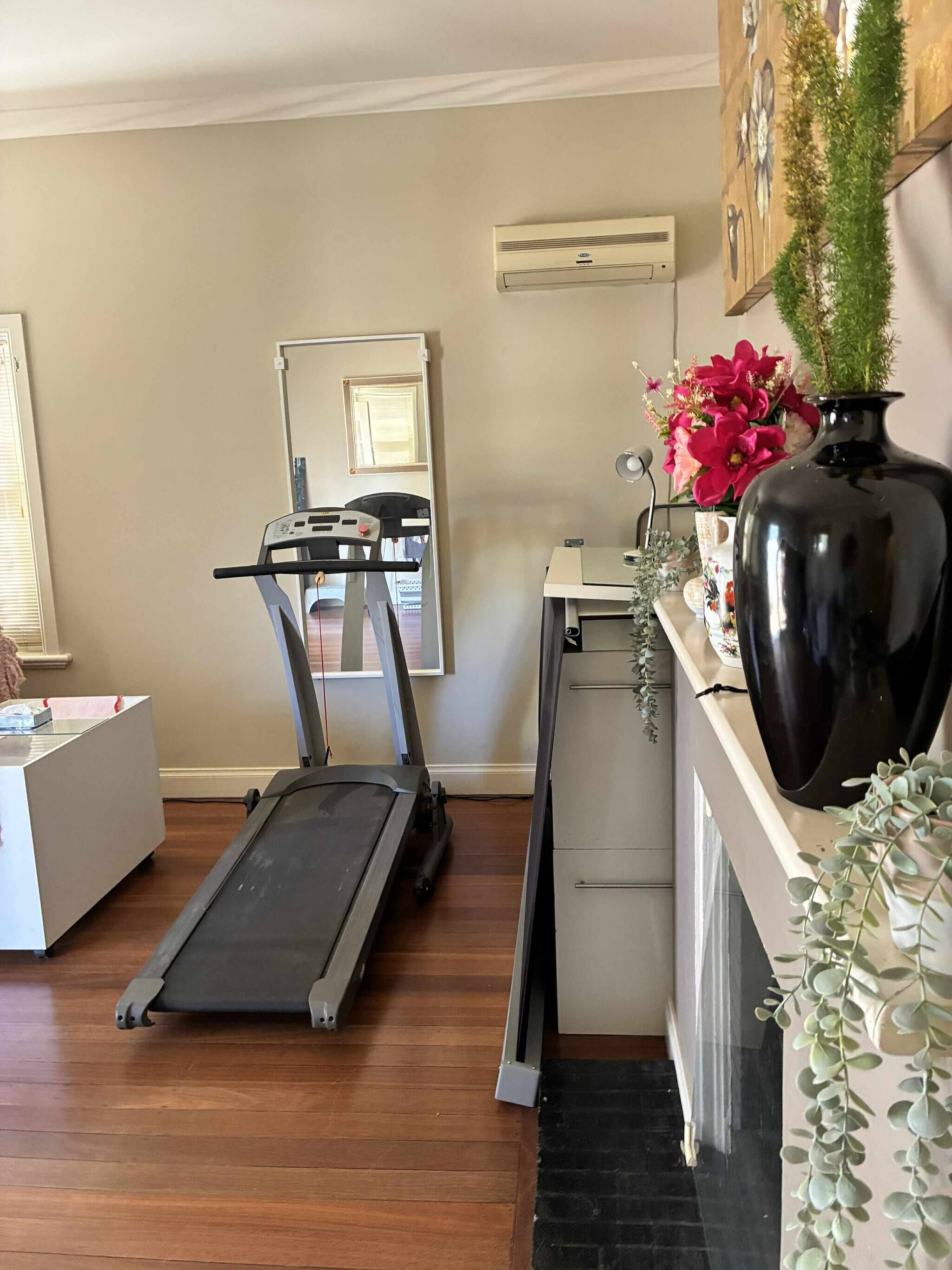 Un gimnasio en casa con una cinta de correr, un escritorio y un jarrón grande con flores.